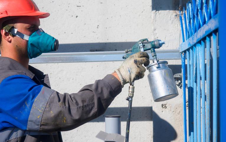 profissional usando máscara, luva e chapéu enquanto utiliza equipamento para pintar portão de azul representando como usar máquina de pintura airless