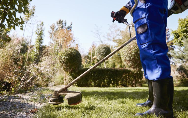 homem com epis azul e preto utilizando roçadeira em local arborizado com grama verde